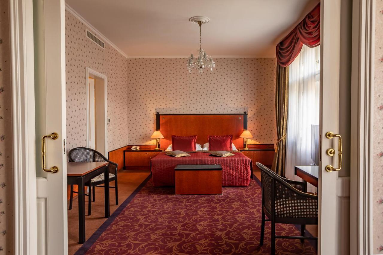 แกรนด์โฮเต็ล พัปป์ Hotel การ์โลวีวารี ห้อง รูปภาพ The photo shows the interior of a hotel room. Upon entering, you can see a spacious area with a plush, red carpet. The room features a large bed with decorative pillows and a wooden headboard, flanked by bedside tables with lamps. In front of the bed