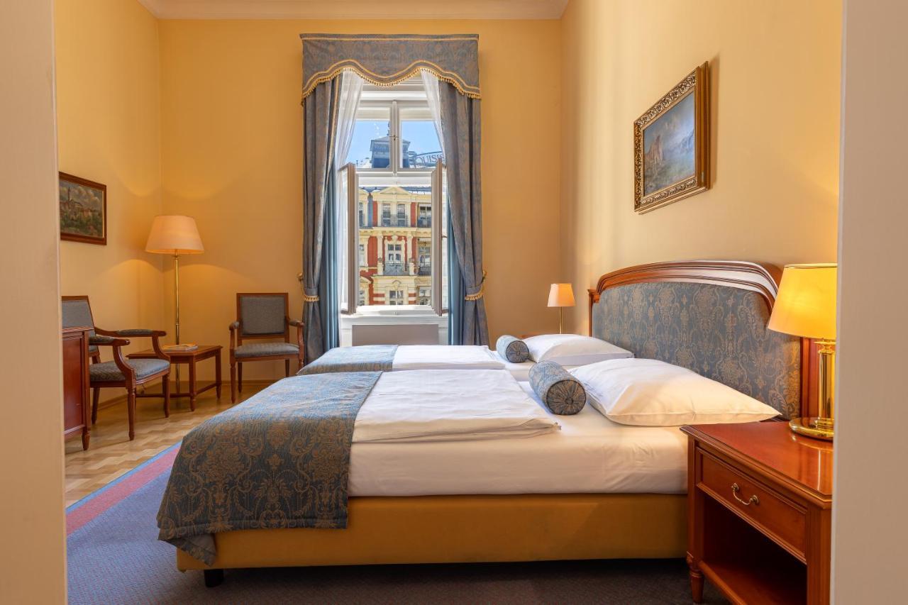 แกรนด์โฮเต็ล พัปป์ Hotel การ์โลวีวารี ภายนอก รูปภาพ The photo depicts a cozy hotel room or bedroom. It features a neatly made bed with a blue and white color scheme, including decorative pillows. On one side of the bed is a small bedside table with a lamp. The room has warm yellow walls and a large wi