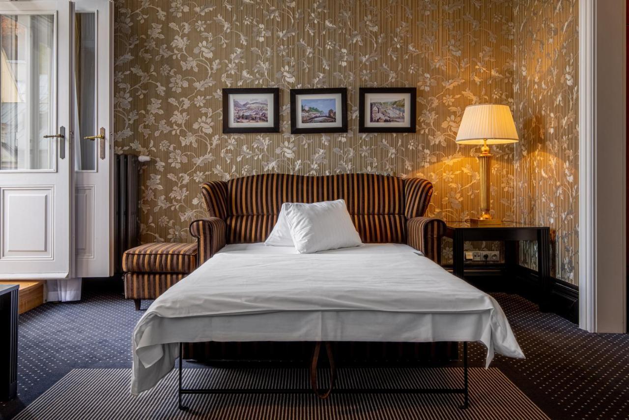 แกรนด์โฮเต็ล พัปป์ Hotel การ์โลวีวารี ภายนอก รูปภาพ The photo depicts a cozy room featuring a striped sofa with a white pillow on it. In front of the sofa, there is a bed or a sleeping surface with a white sheet covering it. The walls are adorned with floral wallpaper, and there are three framed pictu