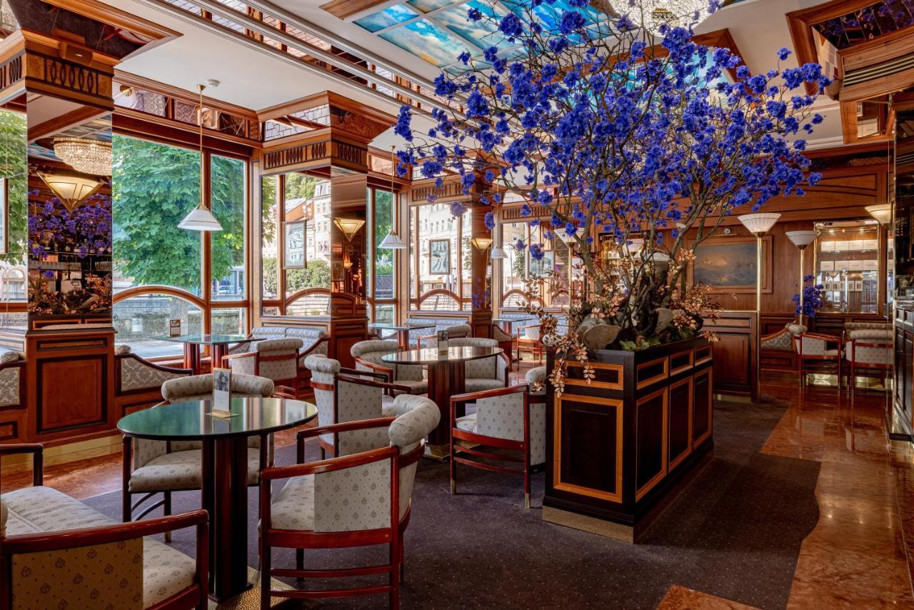 แกรนด์โฮเต็ล พัปป์ Hotel การ์โลวีวารี ภายนอก รูปภาพ The photo shows an elegant interior of a restaurant or café. It features wooden furnishings with intricate details, including circular tables and upholstered chairs. A large floral arrangement, predominantly featuring blue flowers, is placed in the c