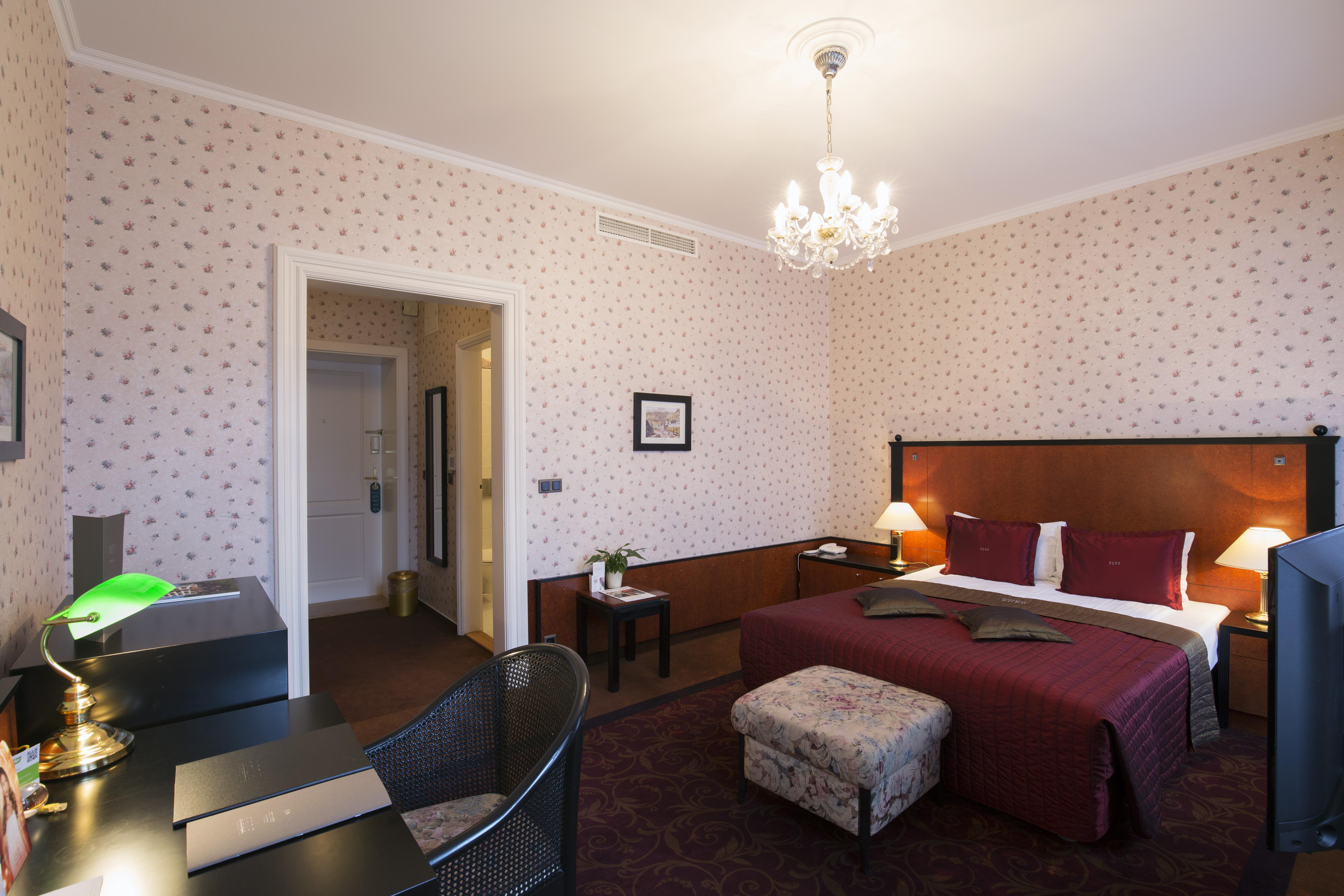 แกรนด์โฮเต็ล พัปป์ Hotel การ์โลวีวารี ภายนอก รูปภาพ The photo depicts a well-furnished hotel room. There is a large bed with a burgundy bedspread, complemented by decorative pillows. The room features soft lighting from a chandelier and lamps on either side of the bed. 

To the left, there is a desk w