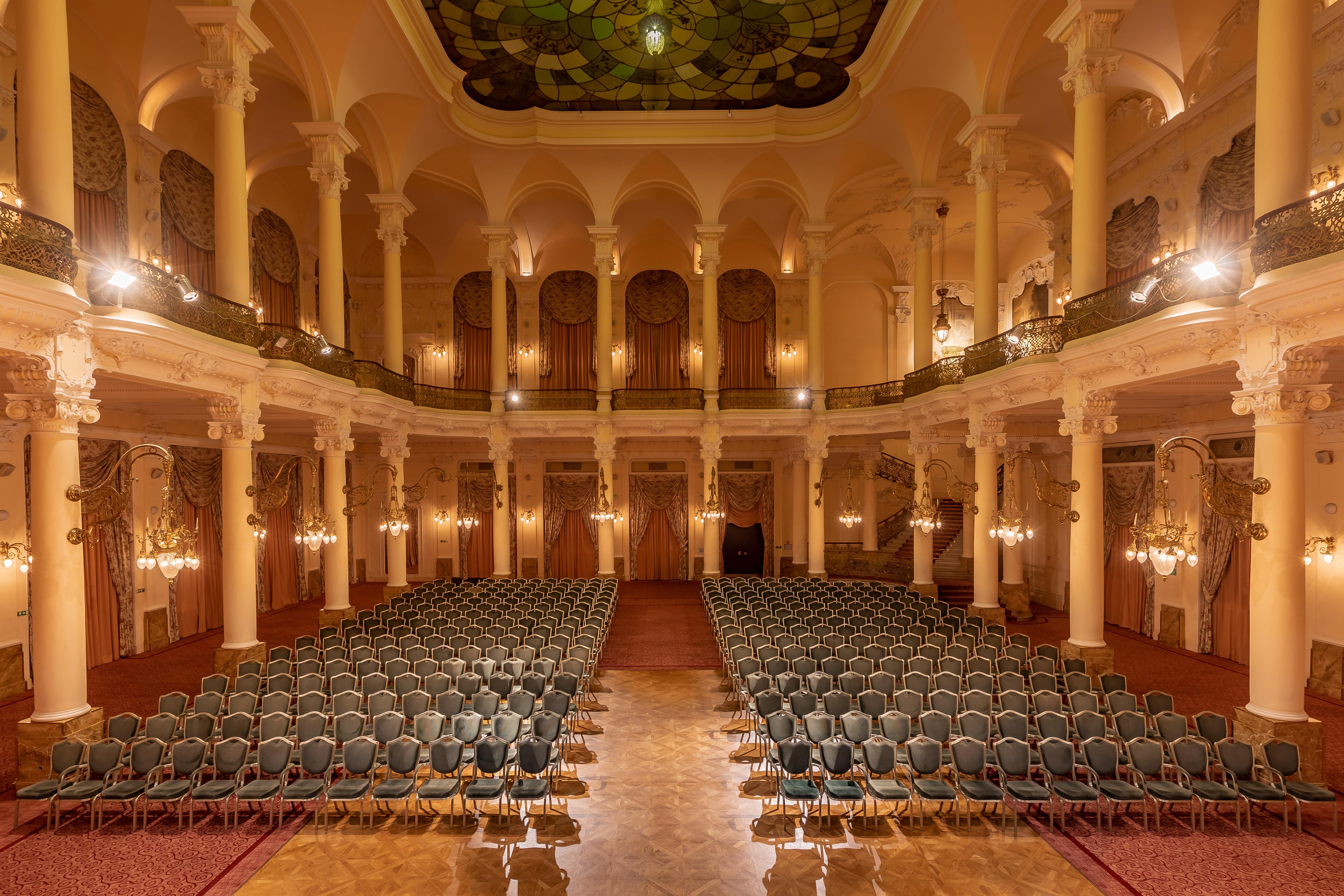 แกรนด์โฮเต็ล พัปป์ Hotel การ์โลวีวารี ภายนอก รูปภาพ The photo shows an elegant, spacious auditorium or hall, designed for events or performances. The interior features high ceilings supported by tall, decorative columns. Rows of green upholstered chairs are neatly arranged, suggesting the space is set