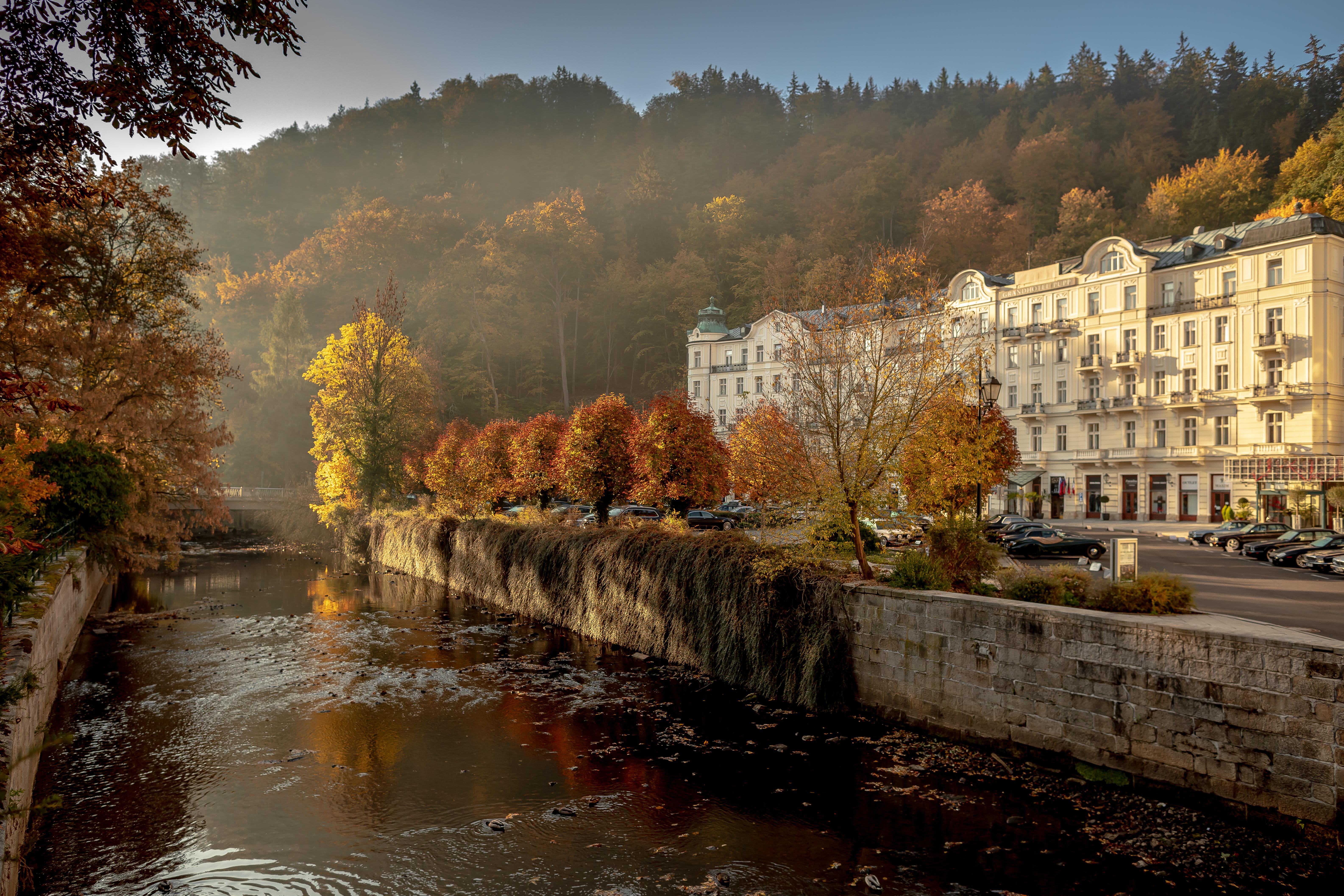 แกรนด์โฮเต็ล พัปป์ Hotel การ์โลวีวารี ภายนอก รูปภาพ The photo depicts a serene riverside scene during autumn. The river flows gently, reflecting the warm hues of the trees along its banks. On one side, there are vibrant orange and yellow trees, showcasing the beautiful fall foliage, while the opposite